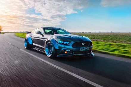 HD PC desktop wallpaper: blue Ford Mustang GT-R muscle car speeding down a country road at sunset — aggressive widebody Ford vehicle with glowing headlights.