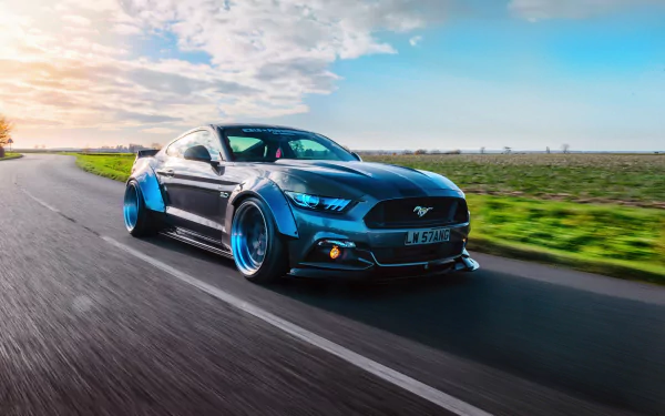HD PC desktop wallpaper: blue Ford Mustang GT-R muscle car speeding down a country road at sunset — aggressive widebody Ford vehicle with glowing headlights.