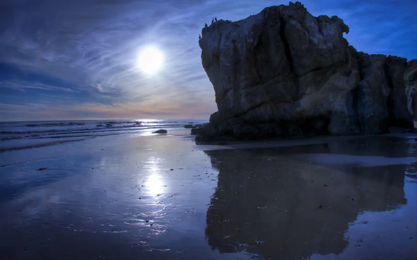 Moonlit night over Malibu beach, California, with ocean waves reflecting the bright moon and a large rock formation silhouetted against the horizon.