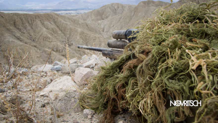 HD PC desktop wallpaper featuring a military sniper camouflaged in a desert landscape, blending into rocky terrain with a clear view of distant sandy hills.