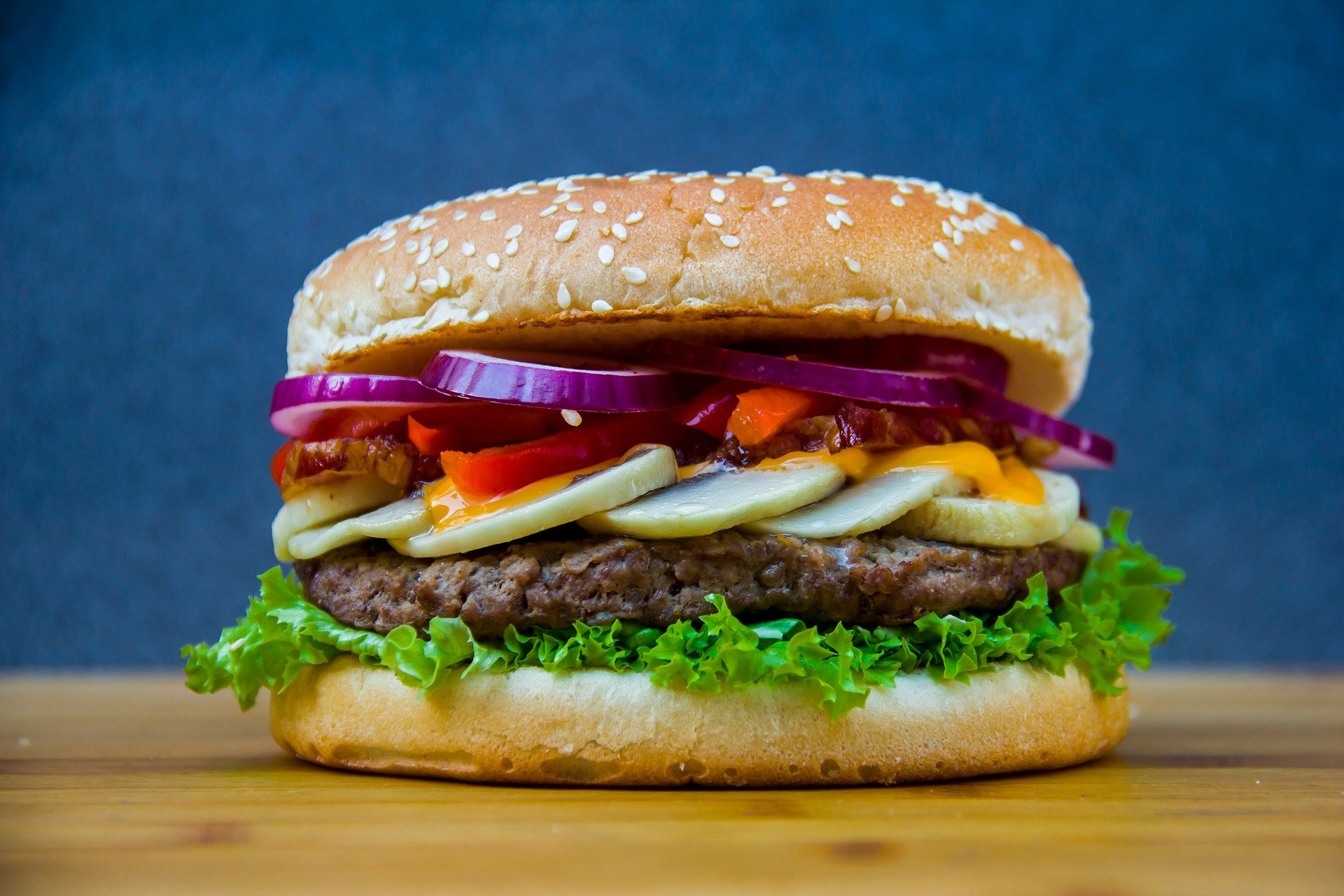 HD PC desktop wallpaper of a juicy hamburger lunch: sesame bun, lettuce, beef patty, melted cheese, red onion and peppers on a wooden surface with blue background.