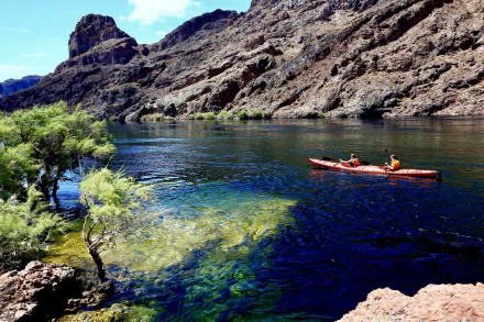 Kayakers paddle a bright red kayak on a clear river winding through an Arizona canyon, surrounded by rocky cliffs and green shrubs under a blue sky.