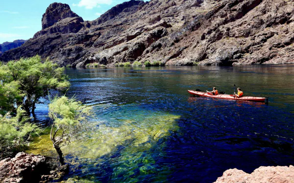 Kayakers paddle a bright red kayak on a clear river winding through an Arizona canyon, surrounded by rocky cliffs and green shrubs under a blue sky.