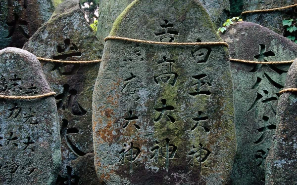 HD desktop wallpaper showing weathered, moss-covered stone statues engraved with Japanese characters, highlighting intricate man-made craftsmanship amidst natural surroundings.