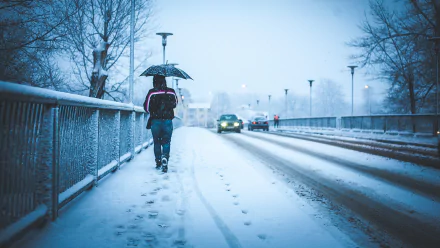 HD winter desktop wallpaper showing a snowy bridge with car headlights, a pedestrian holding an umbrella, and footprints in fresh snow under a soft blue sky.