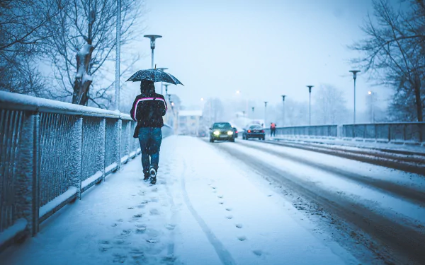 HD winter desktop wallpaper showing a snowy bridge with car headlights, a pedestrian holding an umbrella, and footprints in fresh snow under a soft blue sky.