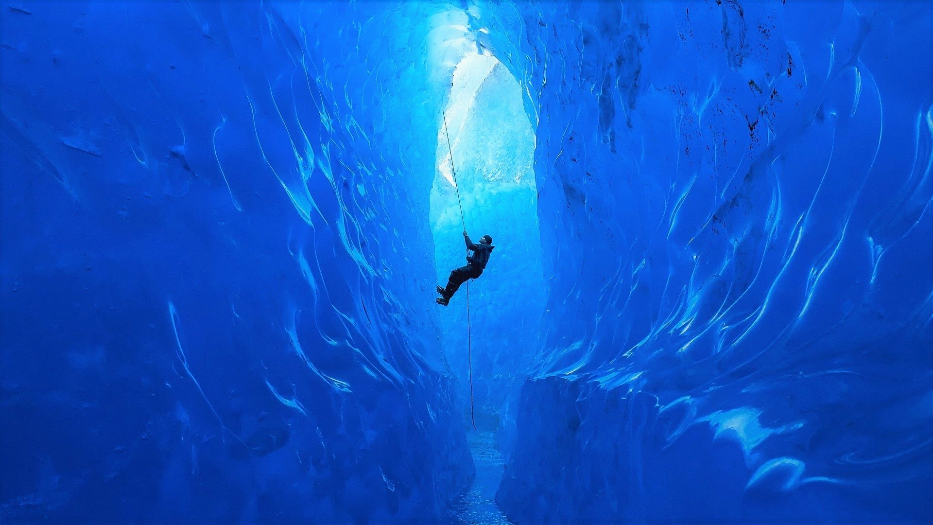 A climber ascends inside a vibrant blue glacier ice cave, showcasing winter ice climbing amid stunning frozen formations.