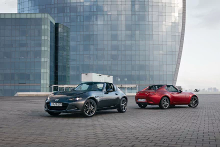 Two Mazda MX-5 cars, one silver and one red, parked side by side in front of a modern glass building.