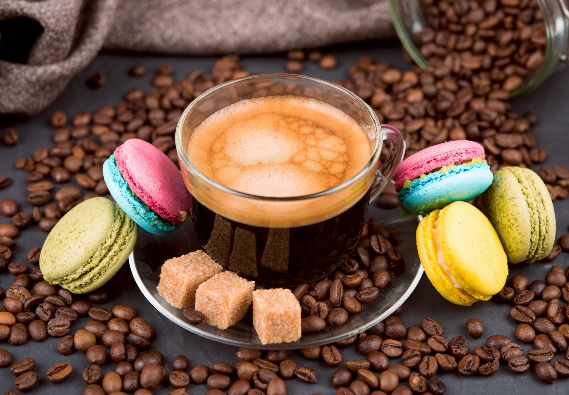 A 4K Ultra HD still life of a clear cup of coffee surrounded by colorful macarons, sugar cubes, and scattered coffee beans on a dark surface.