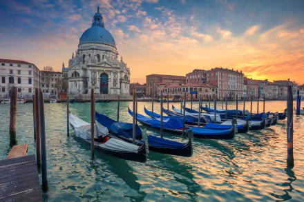 Sunrise over Venice with gondolas docked in the foreground and the dome of historic architecture glowing against the vibrant sky, captured in HD desktop wallpaper quality.