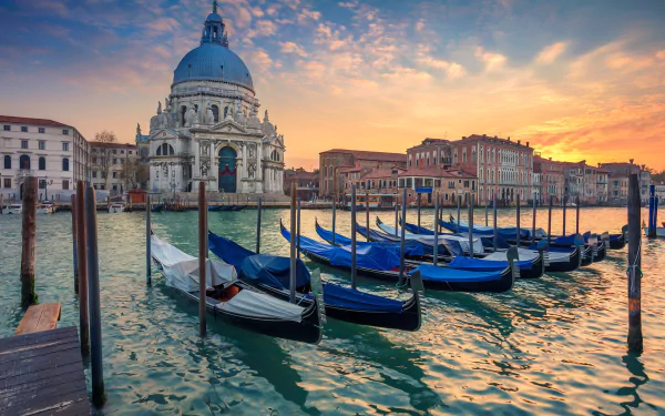 Sunrise over Venice with gondolas docked in the foreground and the dome of historic architecture glowing against the vibrant sky, captured in HD desktop wallpaper quality.