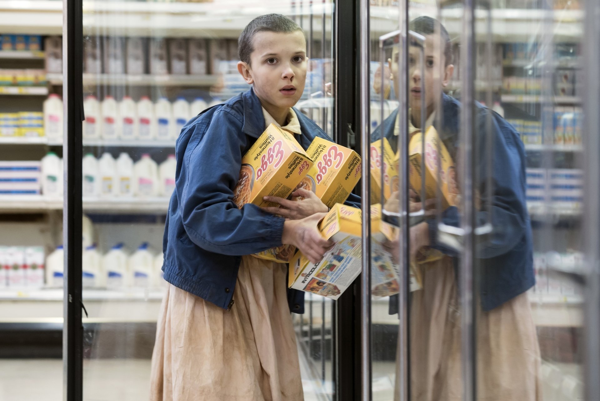 Eleven from Stranger Things, played by Millie Bobby Brown, grabs boxes of Eggo waffles from a supermarket freezer in this 4K Ultra HD TV show wallpaper.