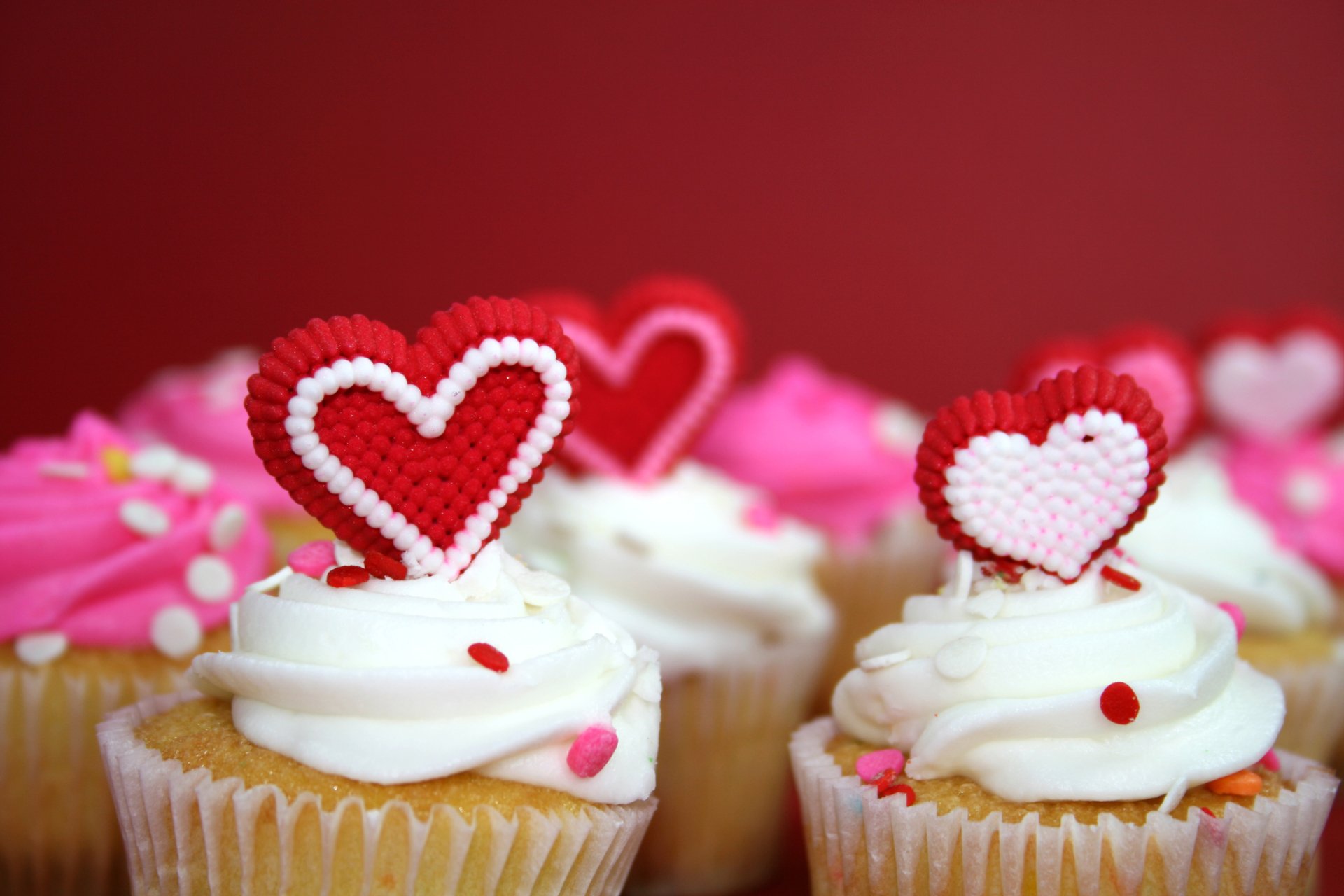 Close-up of cupcakes with white cream and red heart-shaped toppers, featuring vibrant pink and white decorations, styled as a 4K Ultra HD PC desktop wallpaper.