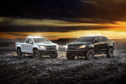 2K Quad HD desktop wallpaper: black and white Chevrolet Colorado pickups on muddy ground in rain beneath a dramatic sunset sky.
