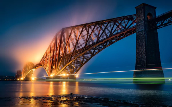 A stunning night view of the Forth Bridge in Edinburgh, Scotland, illuminated with vibrant lights reflecting on the water, creating a captivating man-made landscape.