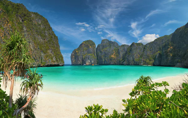 Turquoise waters gently lap the white sandy beach of Phi Phi Island, Thailand, surrounded by dramatic limestone cliffs under a bright blue sky.