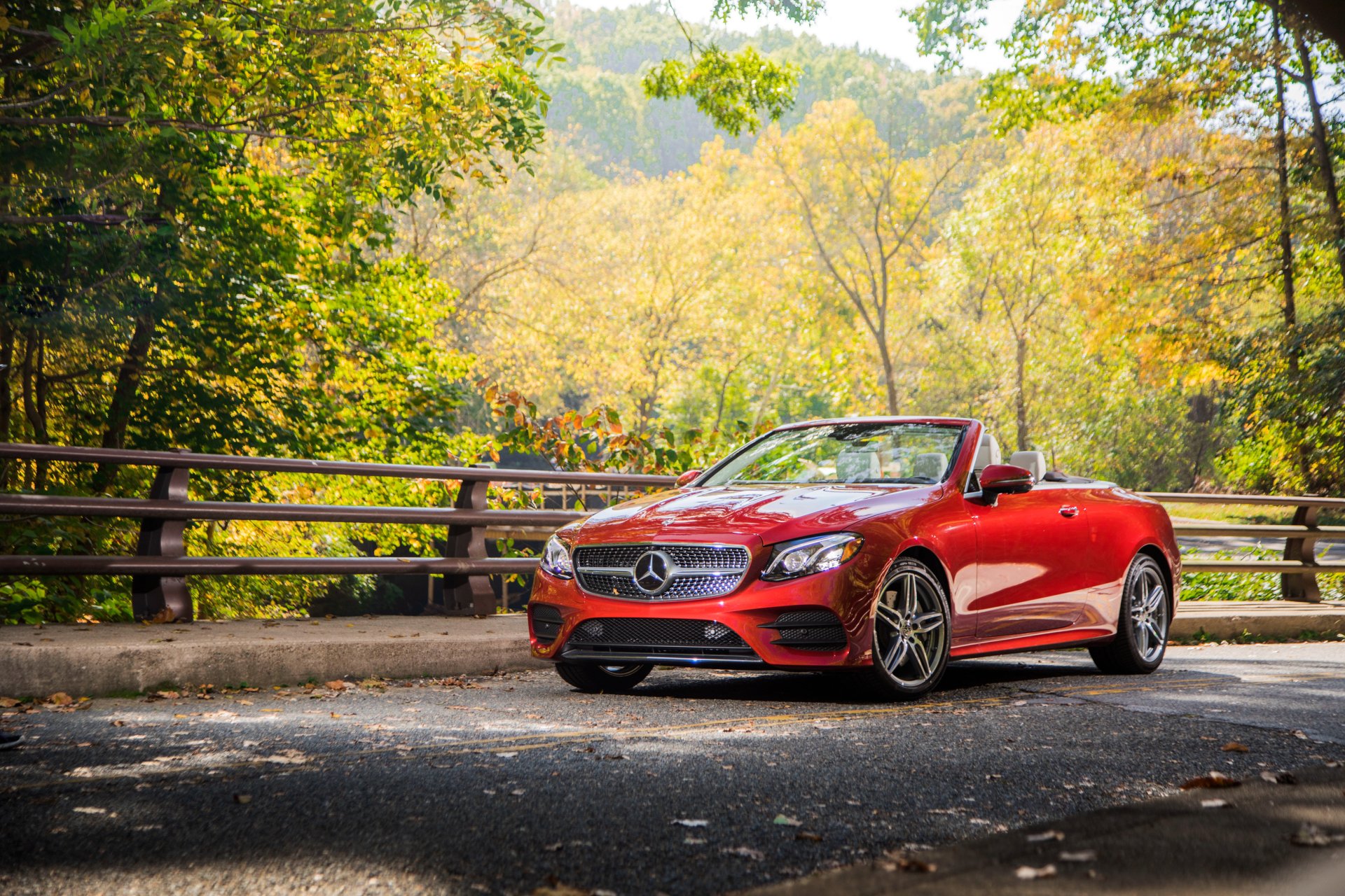 A red Mercedes-Benz E-Class cabriolet parked on a scenic road surrounded by lush green and autumn trees, captured in high definition for a PC desktop wallpaper.