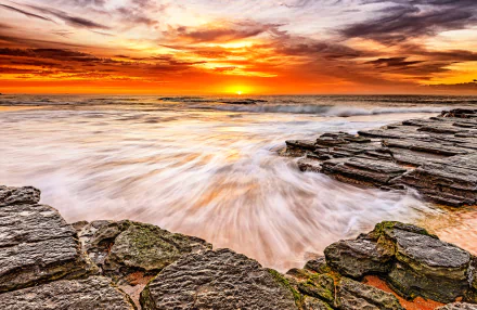HD desktop wallpaper featuring a stunning sunset over the ocean with waves crashing against slate rocks, showcasing the beauty of nature.