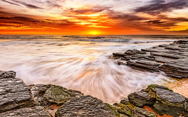 HD desktop wallpaper featuring a stunning sunset over the ocean with waves crashing against slate rocks, showcasing the beauty of nature.