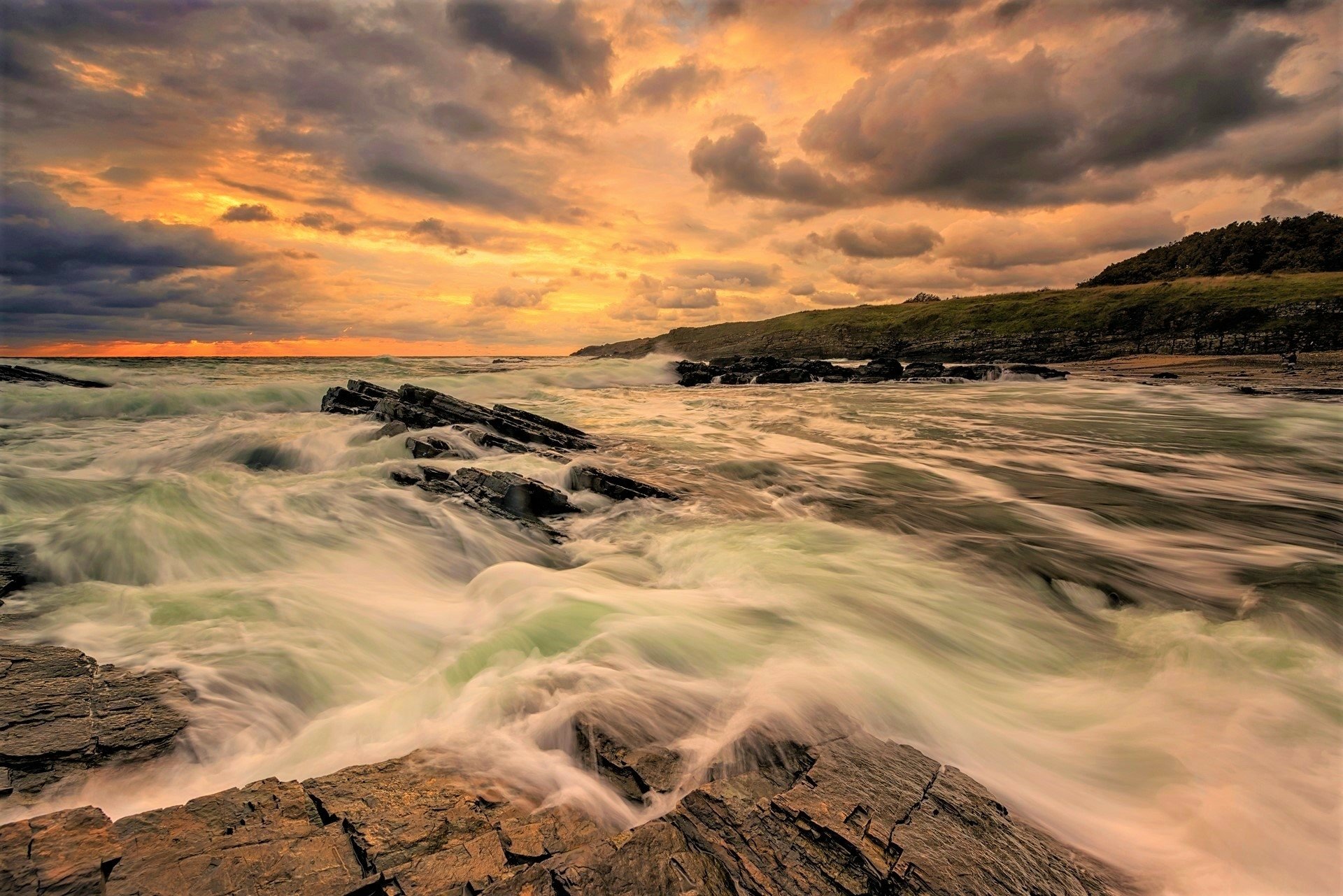 HD desktop wallpaper capturing a dramatic sunset over the ocean with turbulent sea waves, rocky shoreline, and a moody cloudy sky at the horizon.