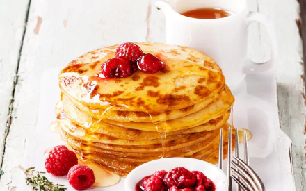 Stack of golden crêpes topped with raspberries and drizzled honey, accompanied by a bowl of berry compote and a white pitcher on a rustic white wooden surface.