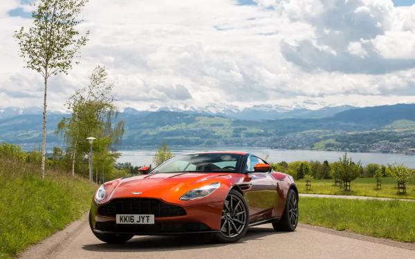 Orange Aston Martin DB11 sports car vehicle parked on a country road with a lake and rolling hills under a cloudy sky — HD PC desktop wallpaper and background.