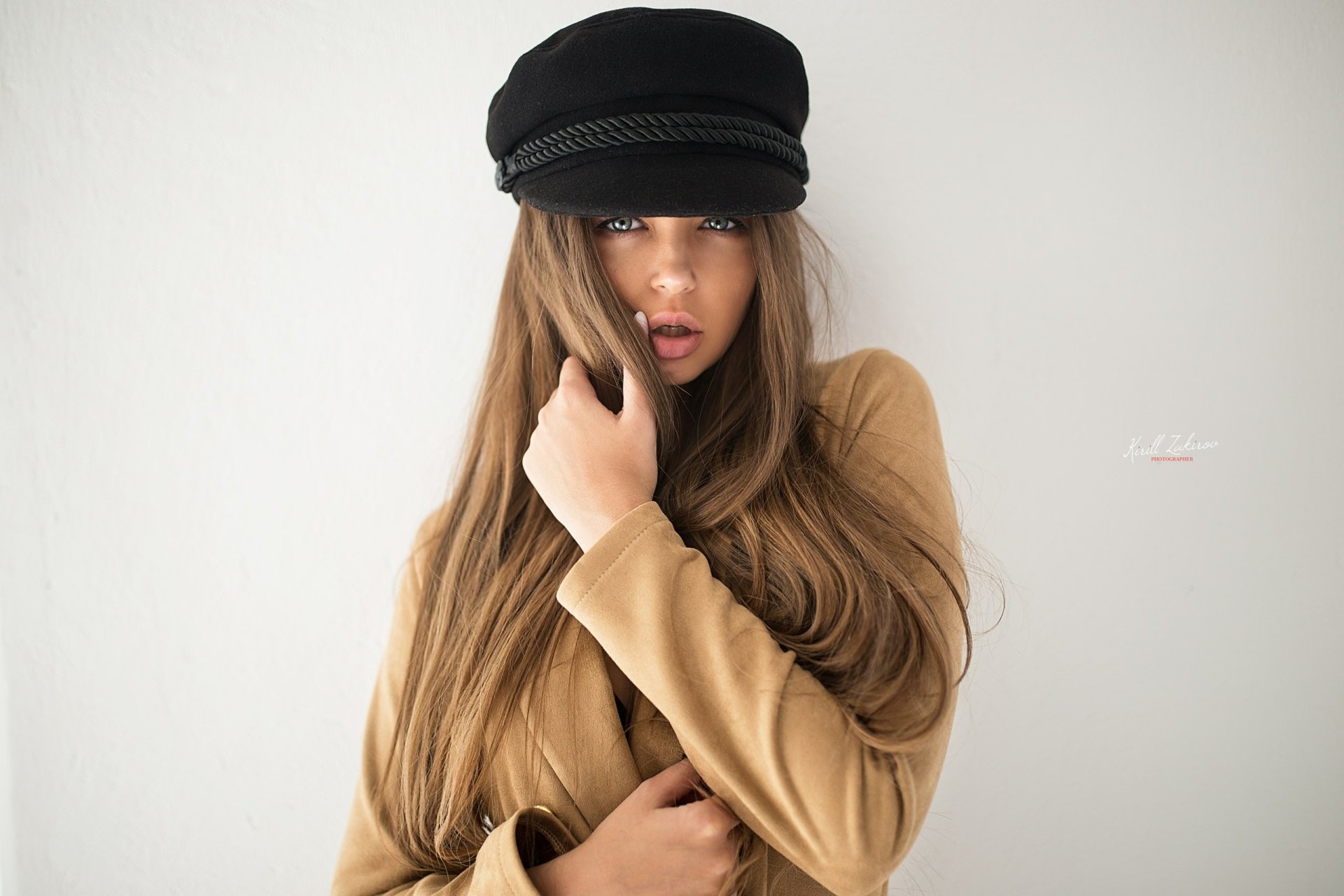 Brunette woman model with long hair and blue eyes wearing a tan coat and black cap, posed against a white background — 2K Quad HD PC desktop wallpaper