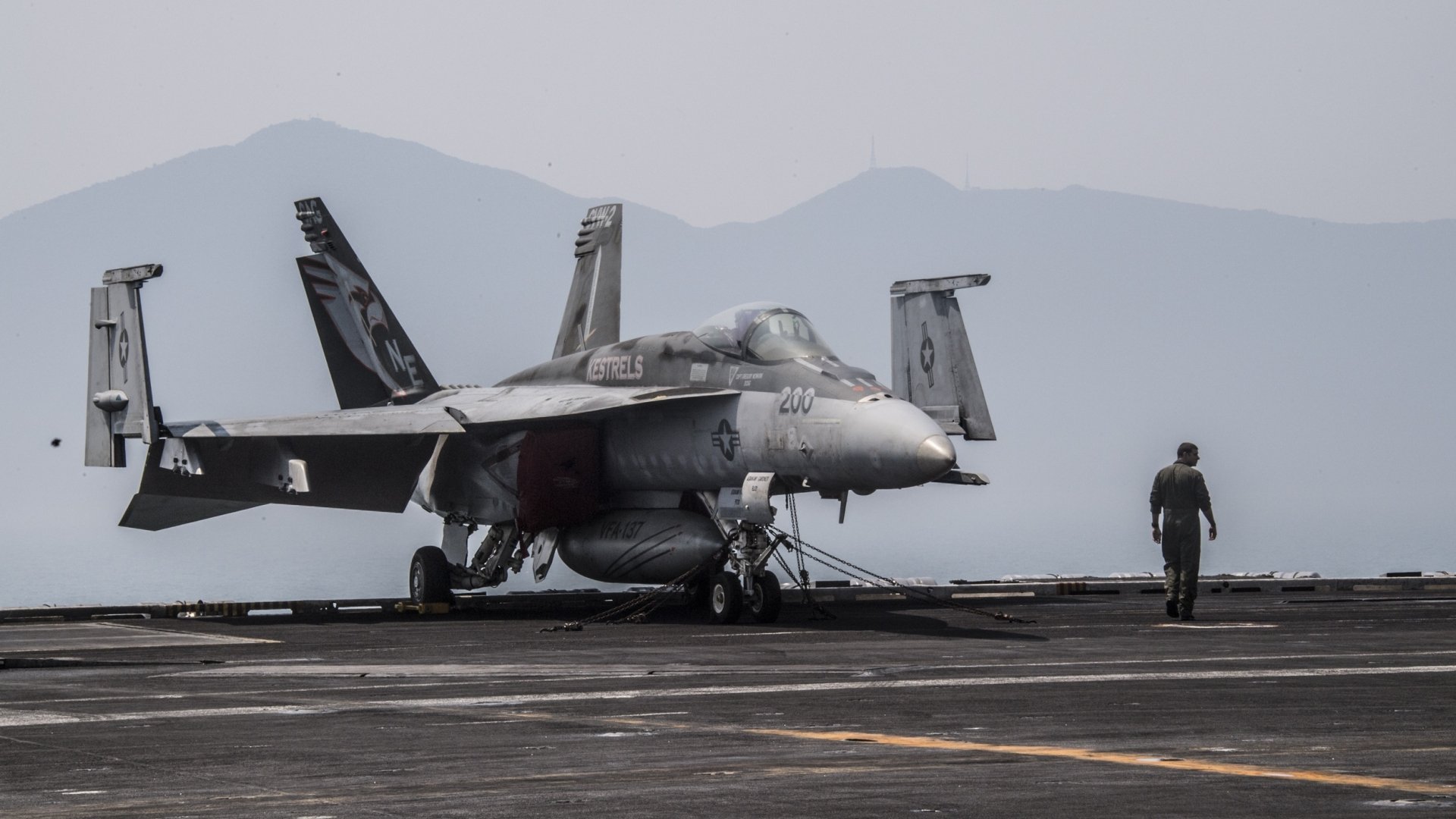 A Boeing F/A-18E/F Super Hornet warplane, a military jet fighter, is stationed on an aircraft carrier deck with a crew member walking nearby, set against a mountainous backdrop.