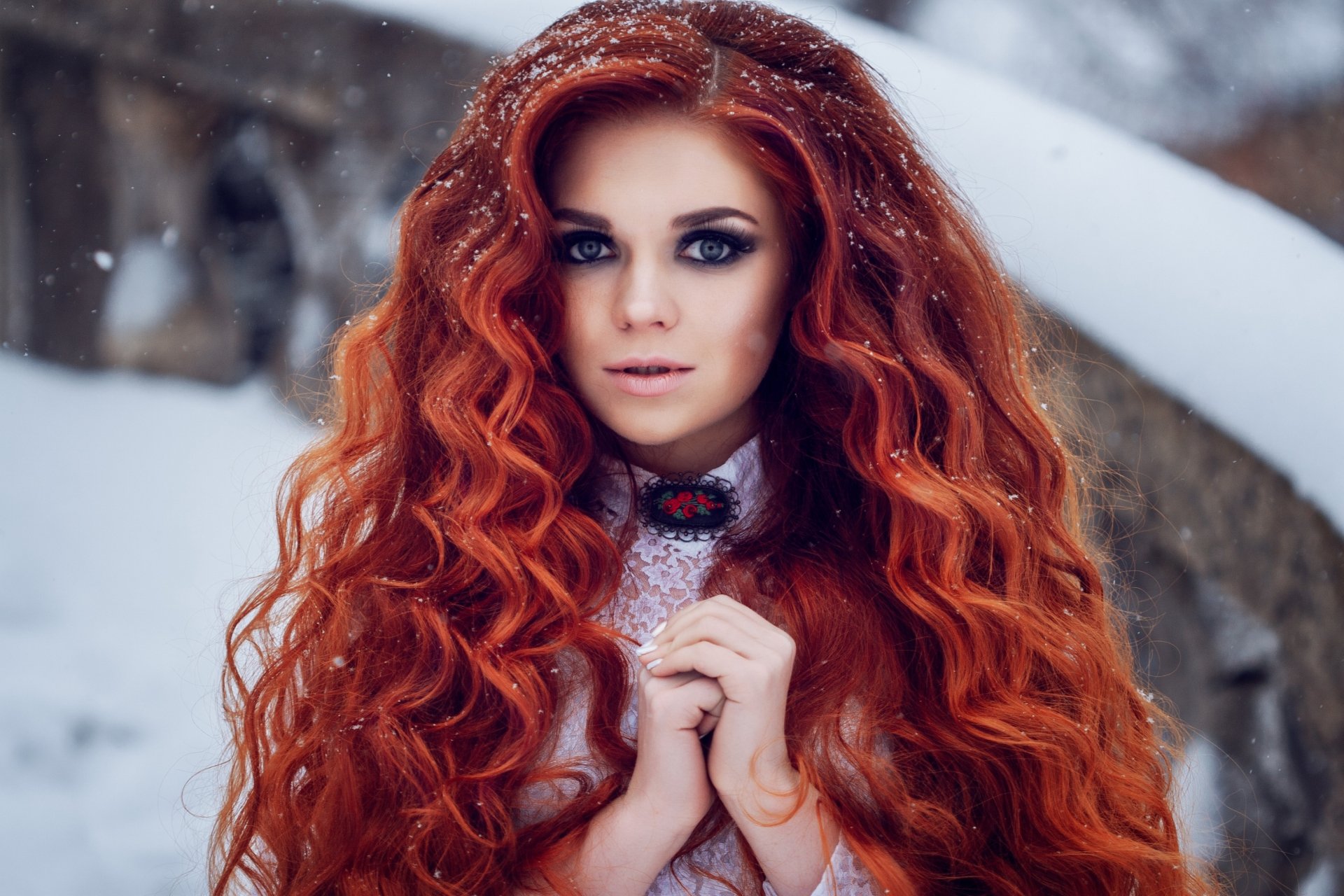 A close-up portrait of a redhead woman with long curly hair and striking blue eyes, captured with depth of field in a snowy outdoor setting, for HD desktop wallpaper.