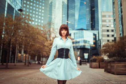 Brunette model in a blue dress with a black corset, framed by shallow depth of field in a glass-towered urban plaza — HD desktop wallpaper.