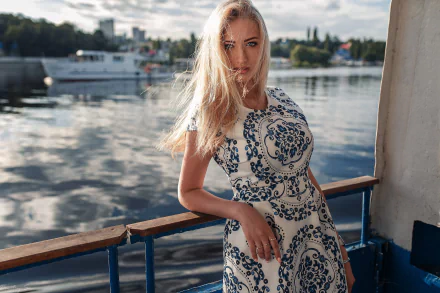Blonde woman model with blue eyes in a patterned dress leans on a boat railing by the water; shallow depth of field, HD PC desktop wallpaper background.