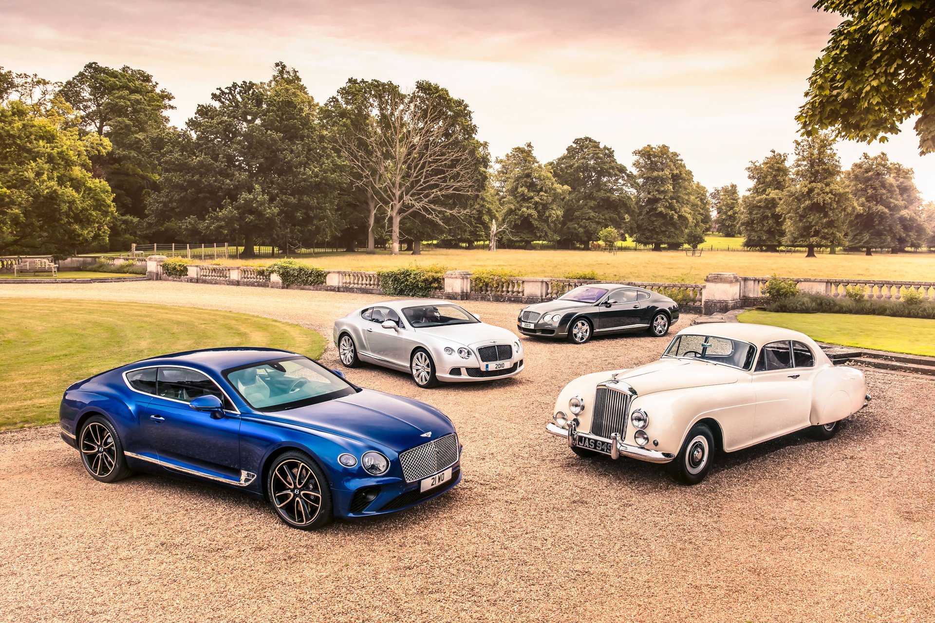 Four luxury Bentley cars, including white and silver models, are parked on a gravel driveway surrounded by green trees, captured in a high-definition desktop wallpaper.