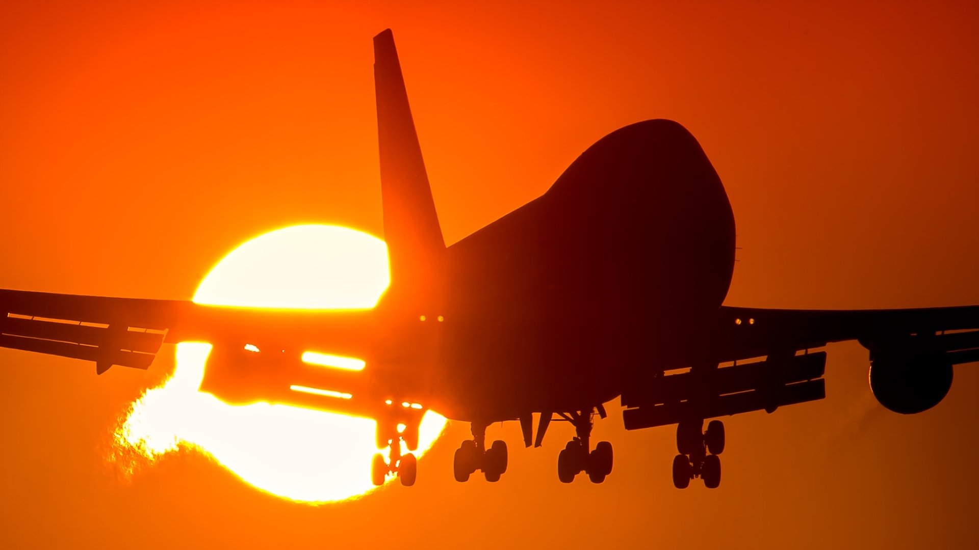 Silhouette of a Boeing 747 passenger aircraft descending toward the glowing sun at sunset — HD PC desktop wallpaper background.