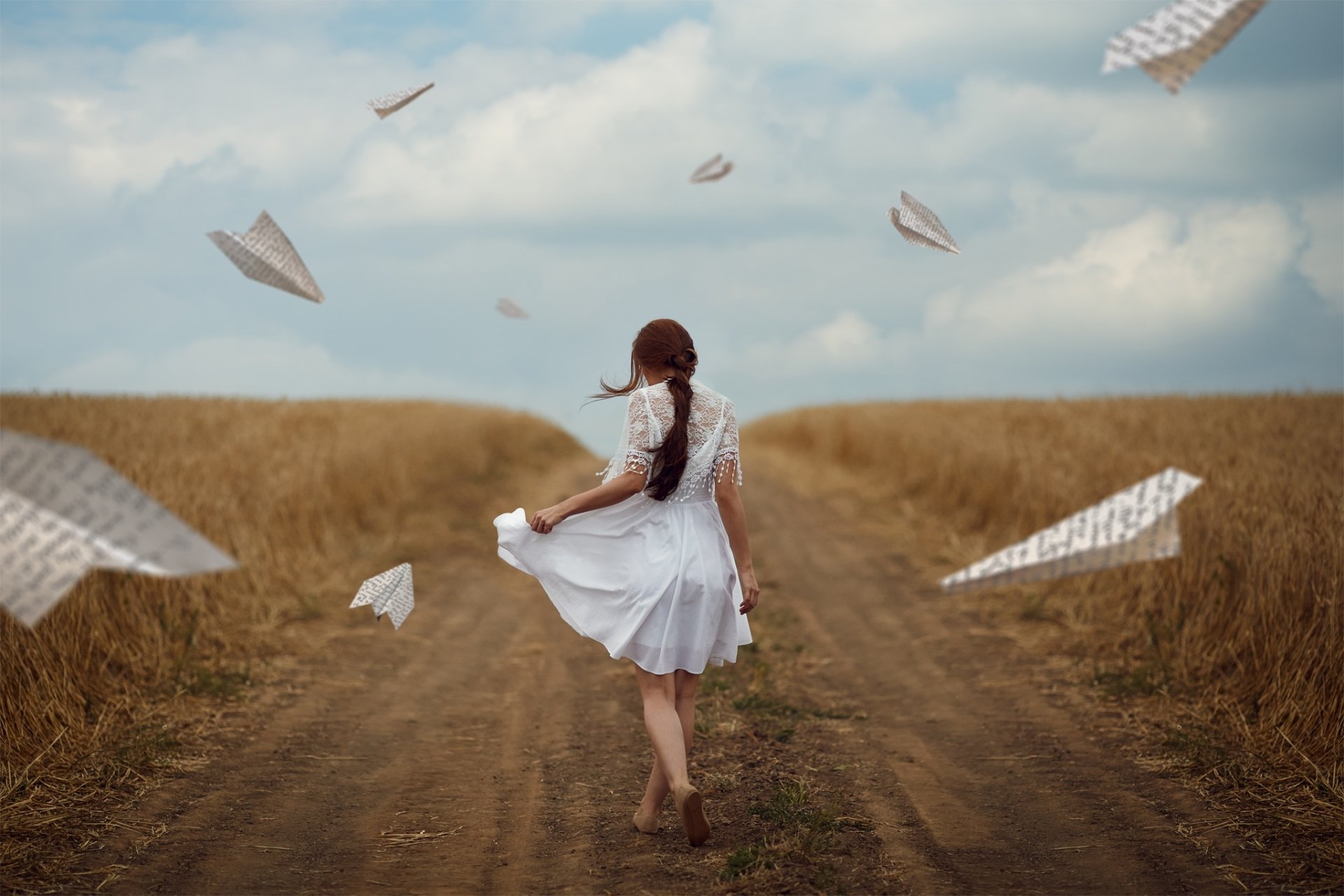 A woman with a braid wearing a white dress walks down a dirt path through a field, surrounded by floating paper planes, captured with artistic depth of field.