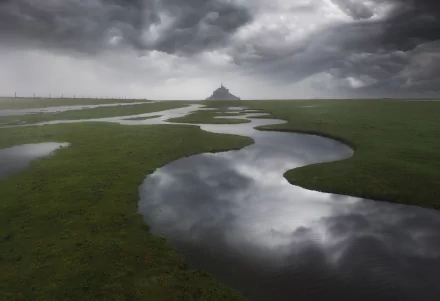 Mont Saint-Michel in France stands under dramatic cloudy skies, reflected in winding water channels across the green landscape in this HD desktop wallpaper.