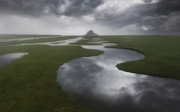 Mont Saint-Michel in France stands under dramatic cloudy skies, reflected in winding water channels across the green landscape in this HD desktop wallpaper.