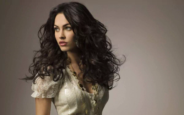 HD desktop wallpaper featuring Megan Fox, with voluminous dark curls and a light-colored, delicate outfit against a neutral background.