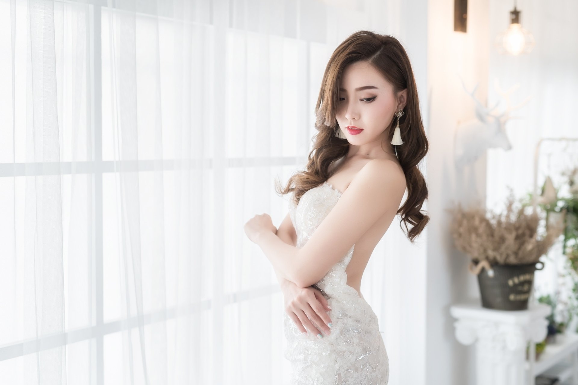 Asian bride with brunette hair in a white wedding dress, wearing earrings and bold lipstick, posing gracefully by a bright window in this HD desktop wallpaper.