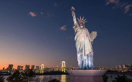 HD desktop wallpaper featuring the man-made Statue of Liberty illuminated at dusk with a city skyline and bridge in the background.