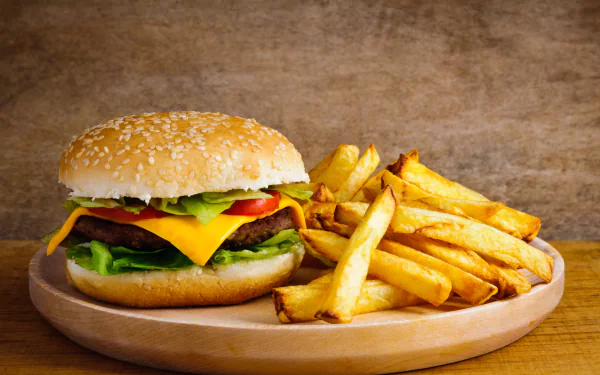 4K Ultra HD image of a cheeseburger with lettuce and tomato, served with a side of golden French fries on a wooden plate against a rustic background.