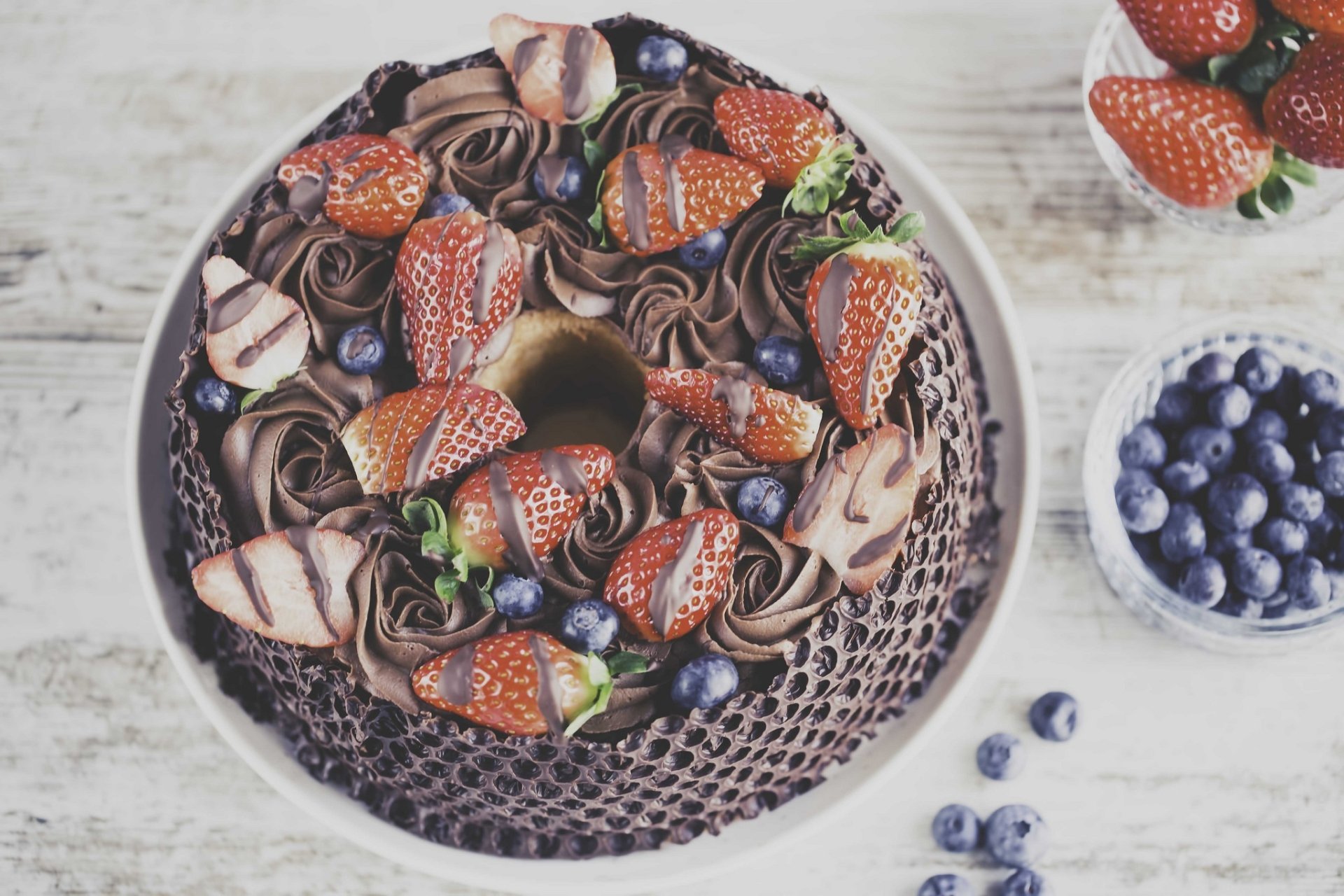 HD PC desktop wallpaper still life: chocolate pastry cake ring topped with strawberries, chocolate rosettes and bowls of blueberries, with a small bluebird accent.