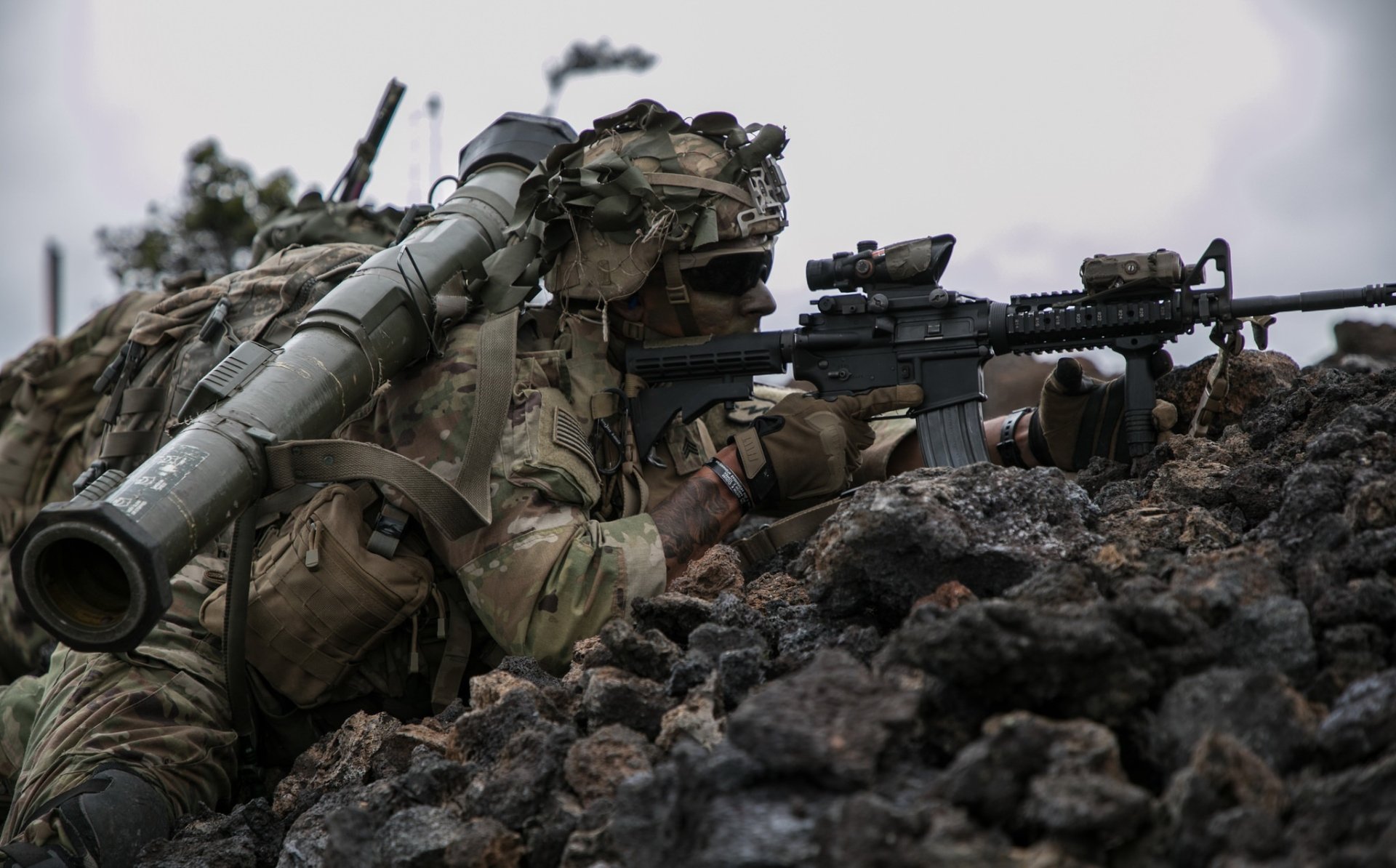 HD military desktop wallpaper showing a soldier in camouflage aiming an M4 Carbine with an AT4 rocket launcher on his back in a rocky battlefield setting.
