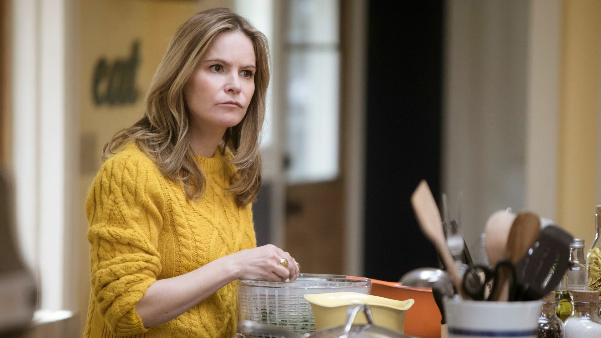 HD desktop wallpaper featuring Jennifer Jason Leigh in a yellow sweater, thoughtfully standing in a kitchen setting.