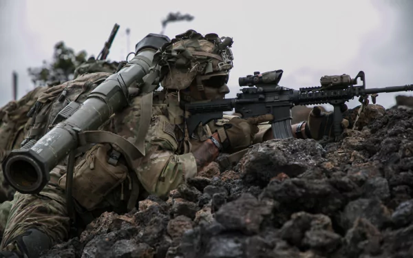 HD military desktop wallpaper showing a soldier in camouflage aiming an M4 Carbine with an AT4 rocket launcher on his back in a rocky battlefield setting.