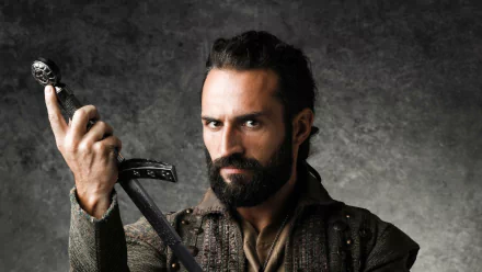 HD desktop wallpaper of a bearded character from Deus Salve o Rei, holding a sword, set against a textured grey background.
