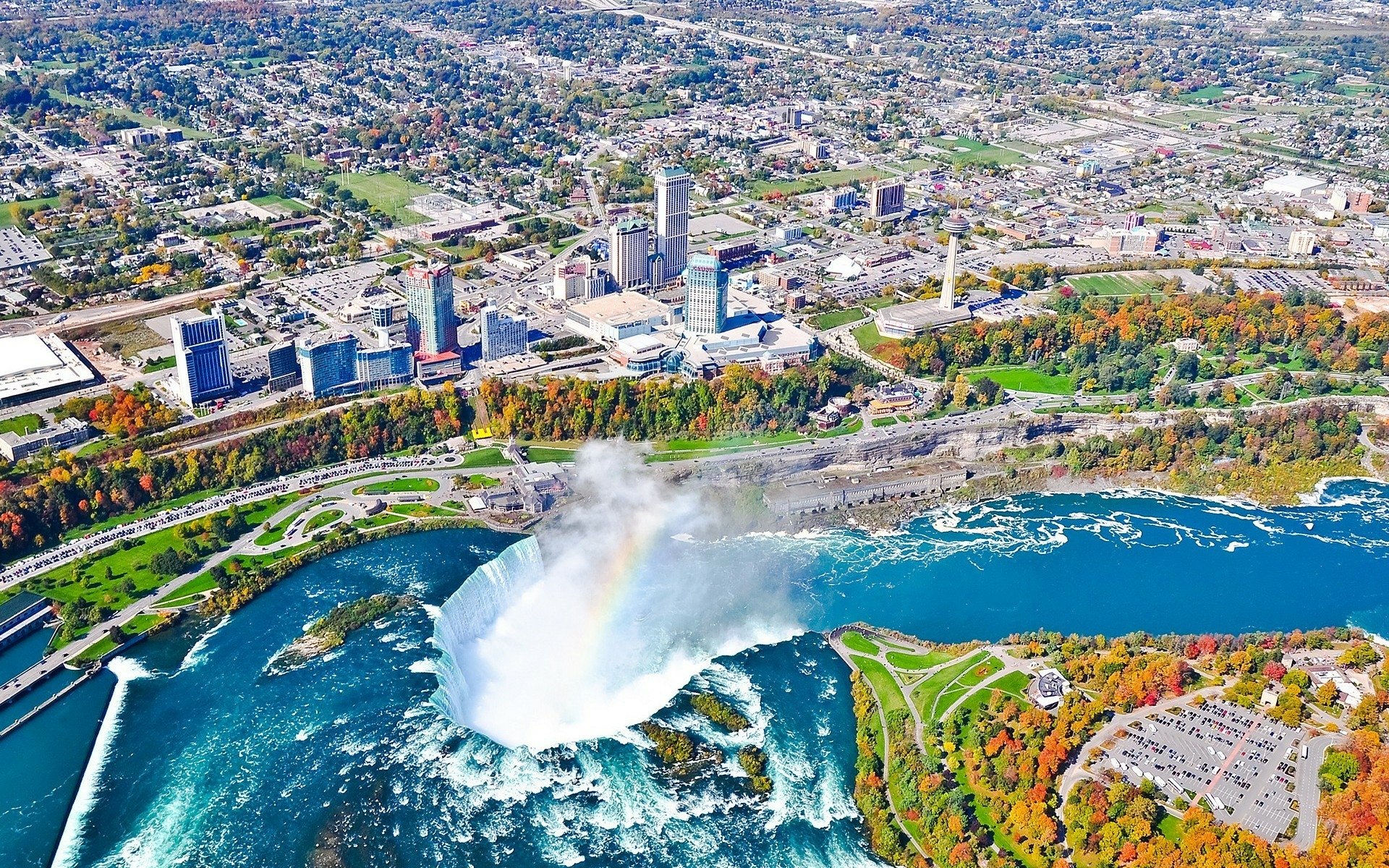 A stunning HD fall view of Niagara Falls with vibrant autumn foliage, the river flowing powerfully between the cityscapes of Canada and the USA.