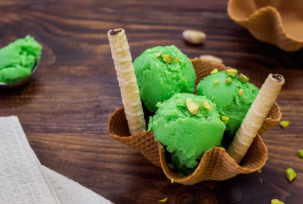 4K Ultra HD PC desktop wallpaper: vibrant green pistachio ice cream scoops in a waffle cone bowl with wafer sticks and chopped nuts on a warm wooden background.