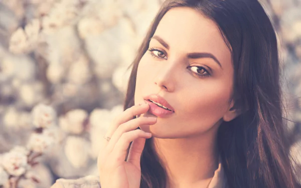 Close-up 5K Ultra HD PC desktop wallpaper of a brunette female model's face with hazel eyes and subtle lipstick against a soft floral background.