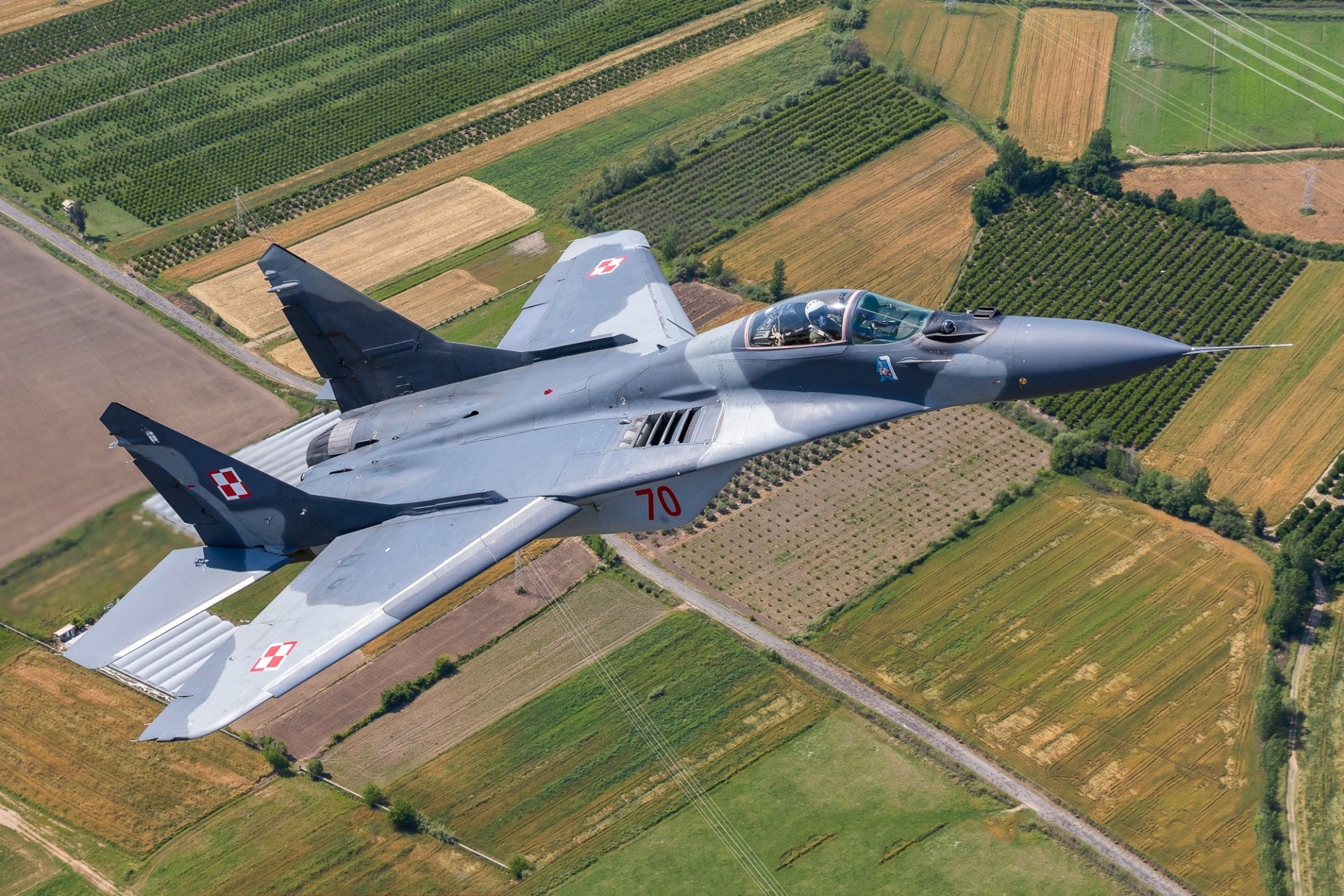 A Mikoyan MiG-29 jet fighter warplane flying over patchwork farmland, captured in high definition as a striking military aircraft desktop wallpaper.