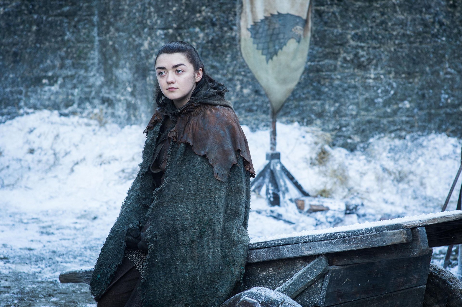 HD desktop wallpaper of Arya Stark (House Stark) from the TV show Game of Thrones, standing cloaked and watchful in a snowy courtyard.